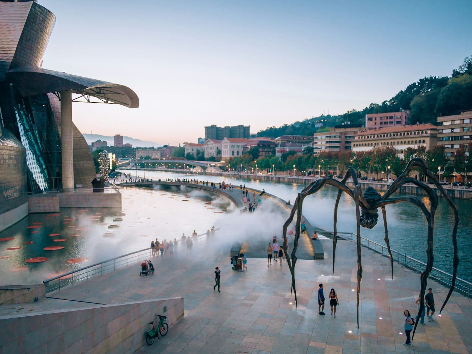 Evening view of Bilbao’s Guggenheim Museum with people walking along the river, city buildings, and iconic spider sculpture under a blue sky.