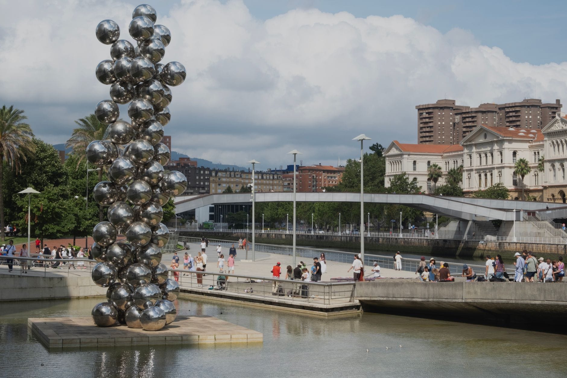 Scena all'aperto a Bilbao con una scultura d'arte moderna di sfere argentate e pedoni vicino al fiume e agli edifici civici.