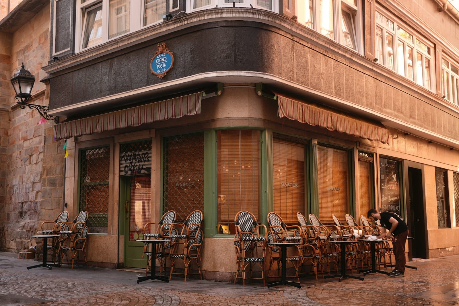 Tavoli all'aperto in un accogliente bar di pintxos angolare su una strada acciottolata nel Casco Viejo di Bilbao, con illuminazione calda e una persona che sistema le sedie.