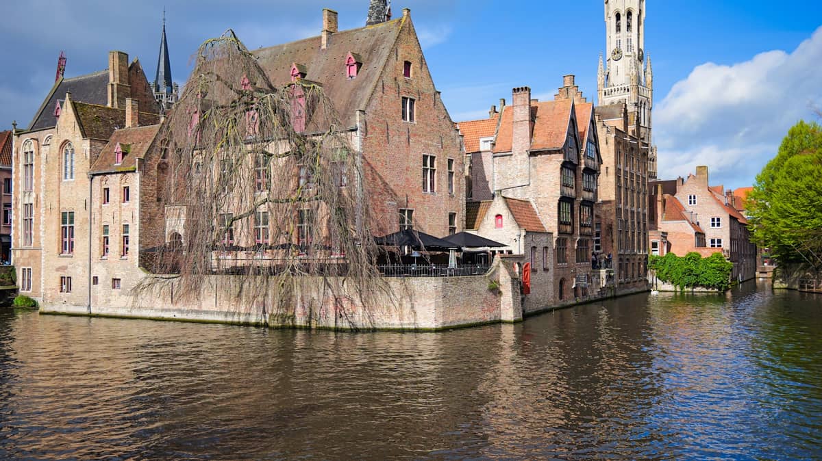 Classic Bruges canal view with medieval brick buildings, the Belfry tower in the background, and water reflecting the sky and historic architecture.