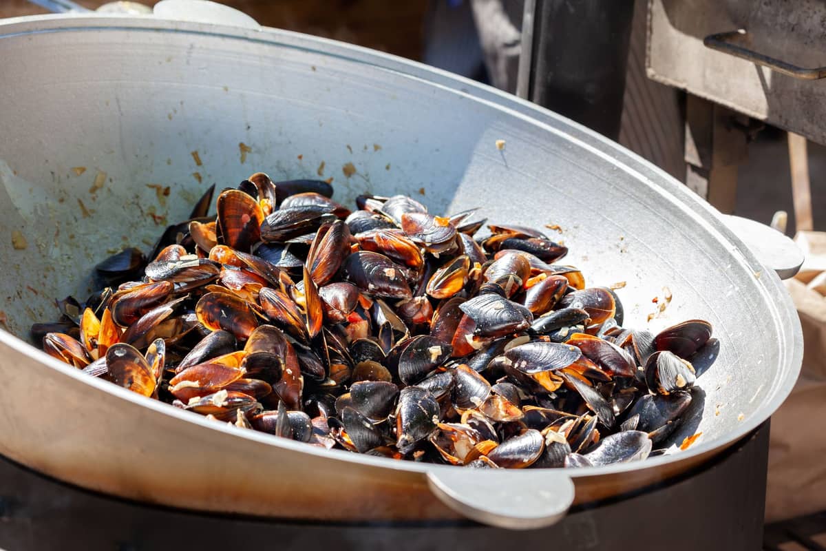 Large metal pan filled with freshly steamed mussels, typical Belgian seafood dish, being cooked outdoors at a food market.