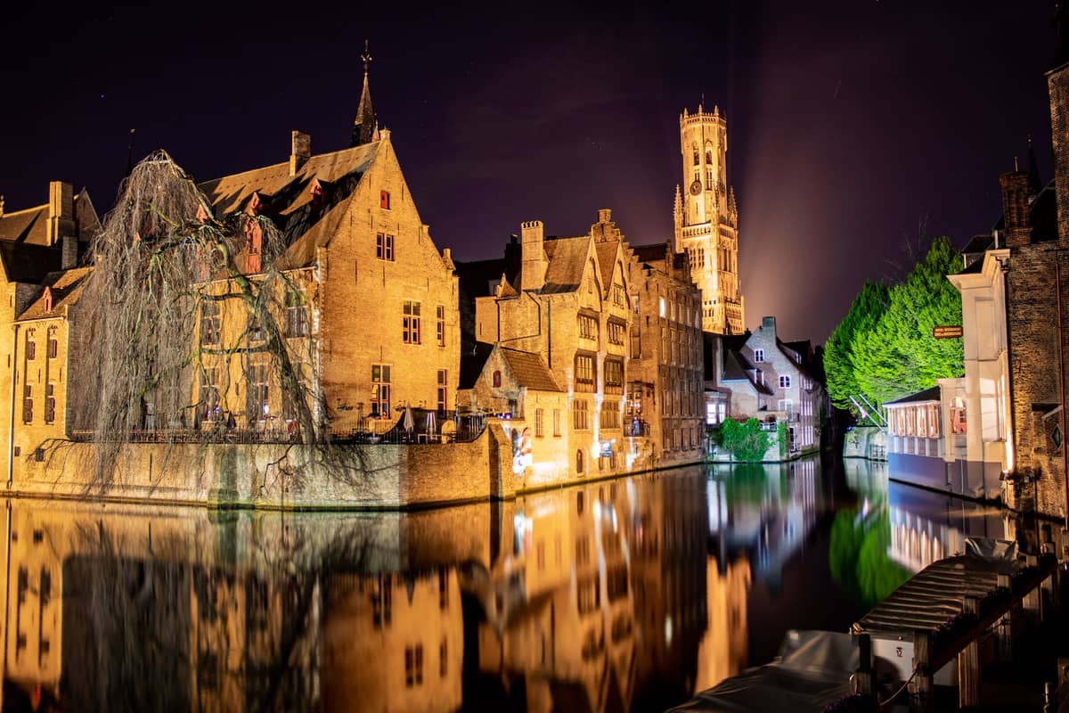 Nighttime view of Bruges' Rozenhoedkaai with illuminated medieval buildings reflected in the canal, empty of crowds, and the Belfry tower in the background.