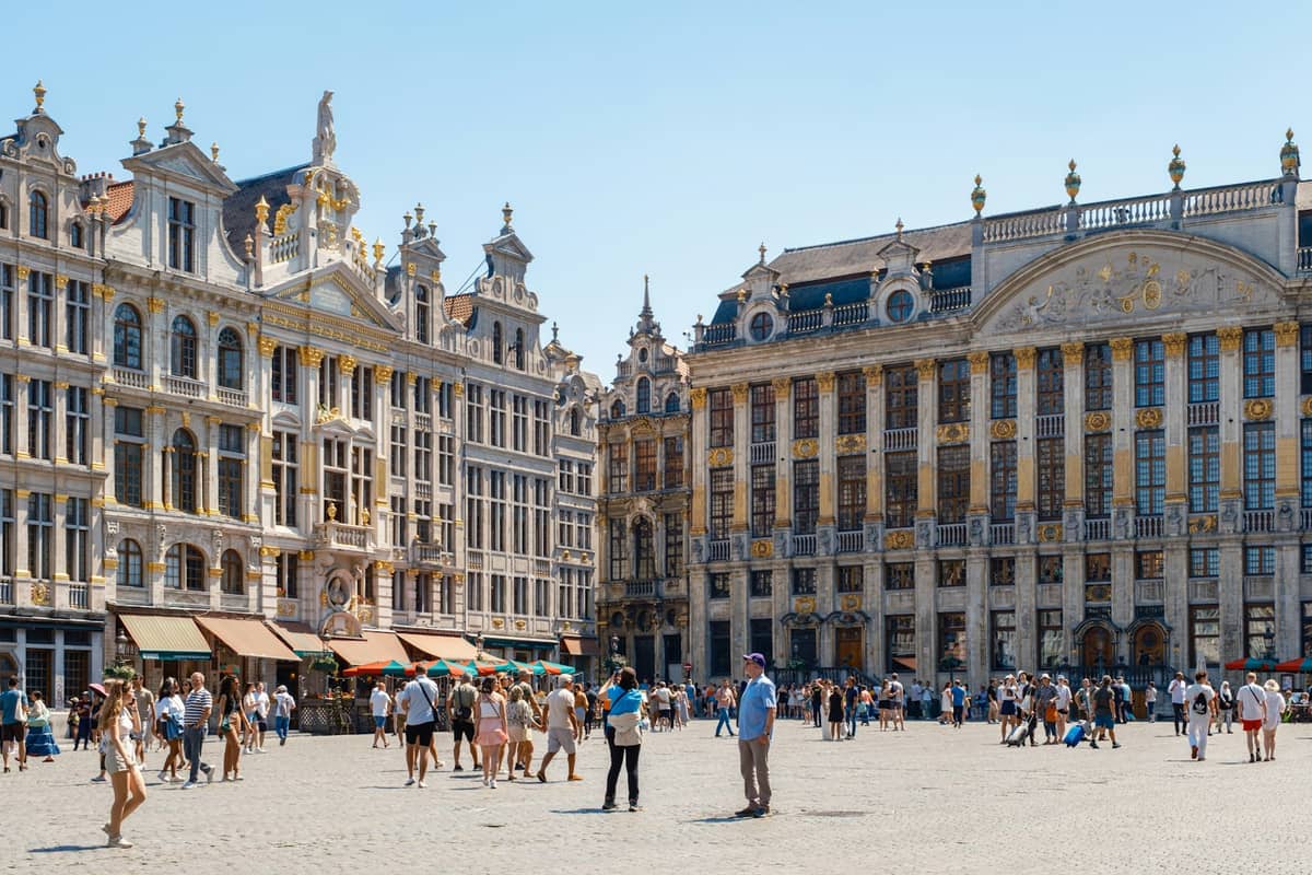 Vista ampla da Grand-Place de Bruxelas com as ornamentadas casas das guildas, pessoas caminhando pela famosa praça histórica e céu azul ao fundo.