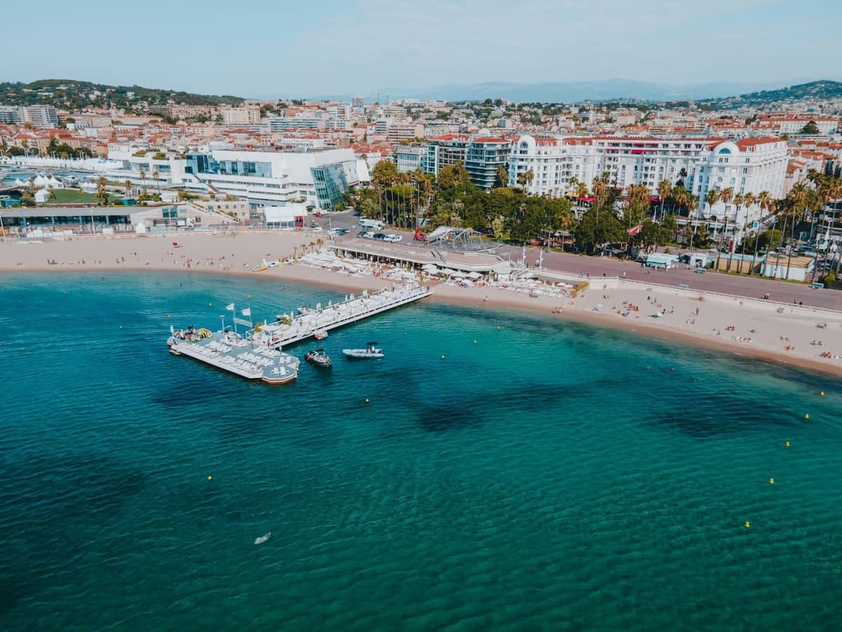 Widok z lotu ptaka na plażę w Cannes, molo, turkusową wodę, łodzie i budynki miasta w tle w pogodny dzień.