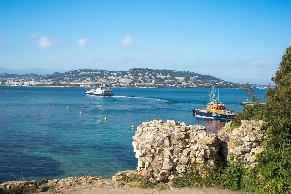 Promy na wodzie z Cannes i wybrzeżem Lazurowego Wybrzeża w tle, widziane z Wysp Lérins z kamiennymi ruinami na pierwszym planie.