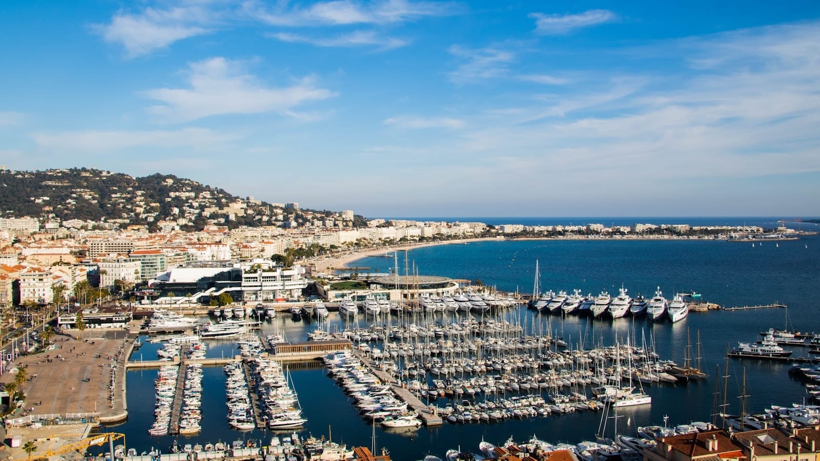 Vista panorâmica deslumbrante do porto de Cannes com iates, orla curva, prédios da cidade e colinas ao fundo sob um céu azul intenso.