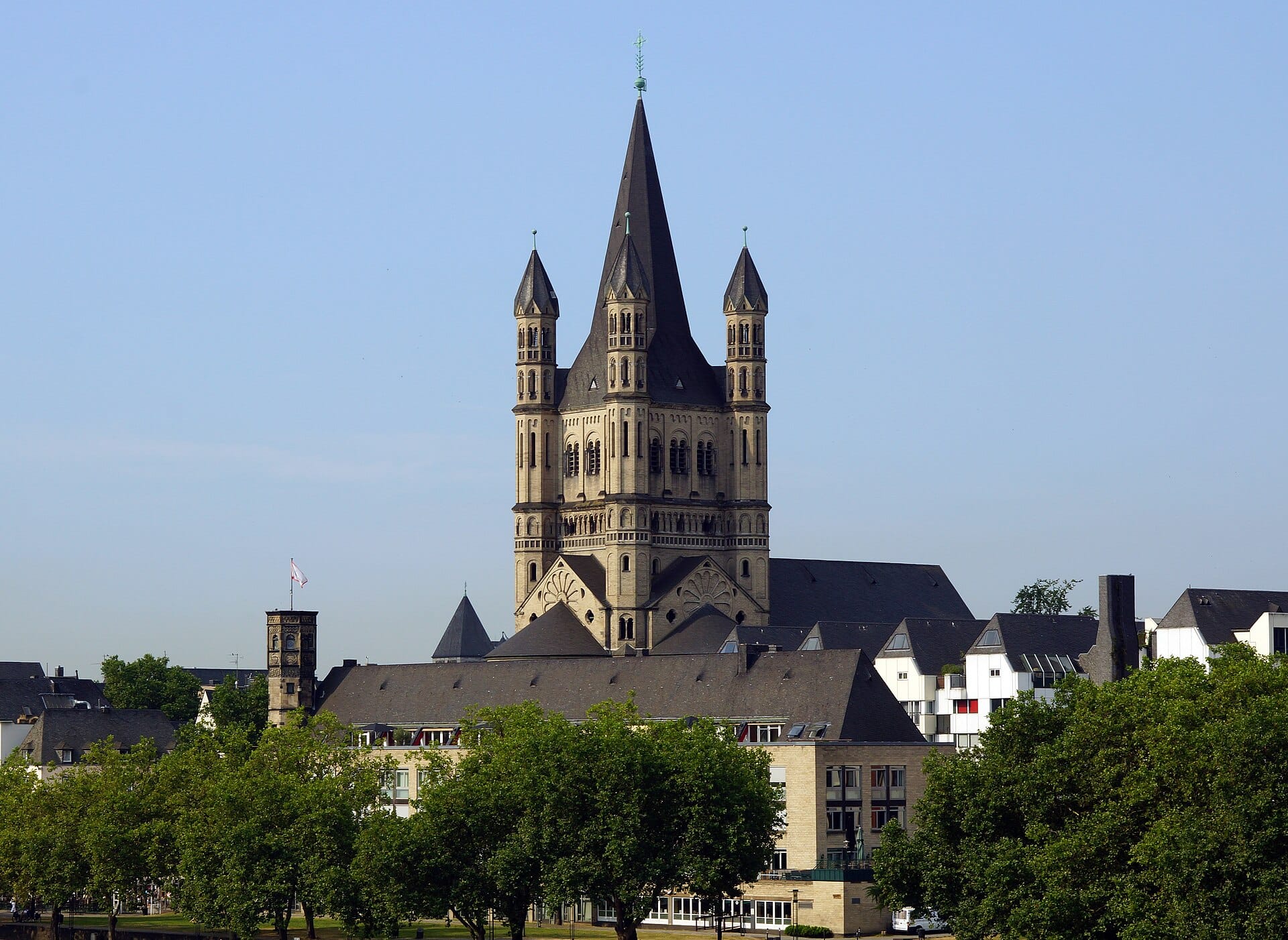 โบสถ์ Groß St. Martin ในเมือง Cologne ตั้งตระหง่านเหนือย่านเมืองเก่า ล้อมรอบด้วยต้นไม้และอาคารแบบดั้งเดิมภายใต้ท้องฟ้าสีฟ้าใส