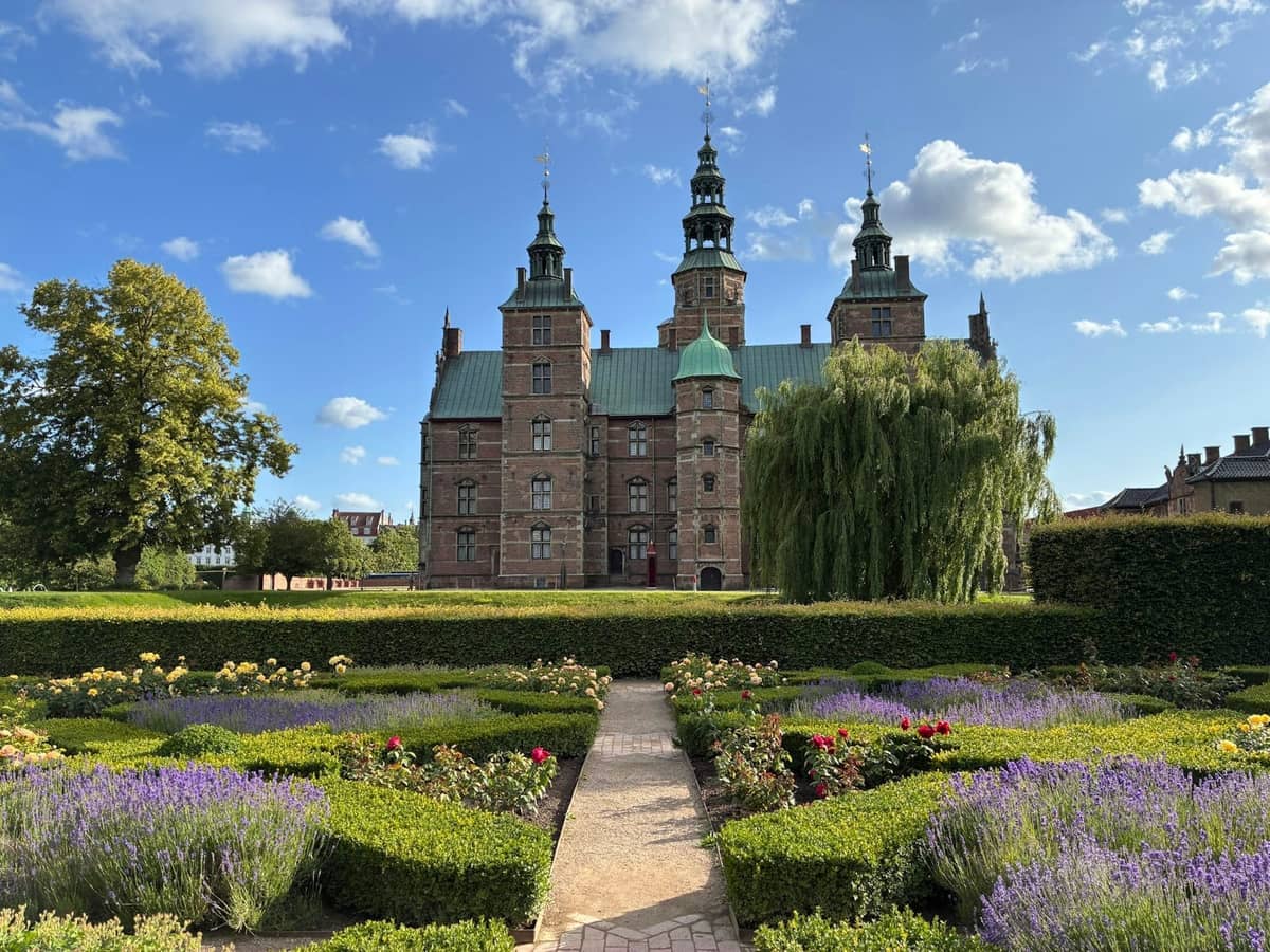 Zamek Rosenborg w Kopenhadze w słoneczny dzień, widziany z bujnego ogrodu z kolorowymi kwiatami i przystrzyżonymi żywopłotami.