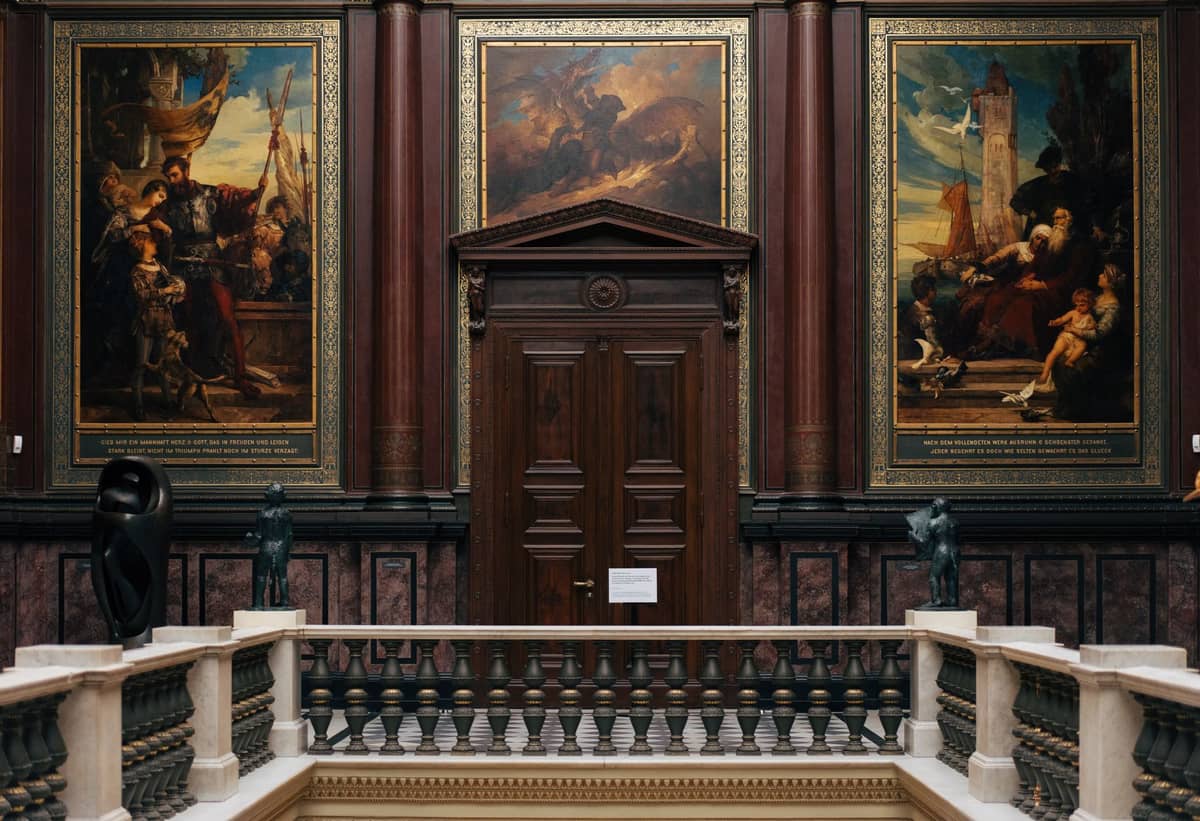 Interior of an art museum with grand staircase and large classical paintings on the wall in an ornate hall.