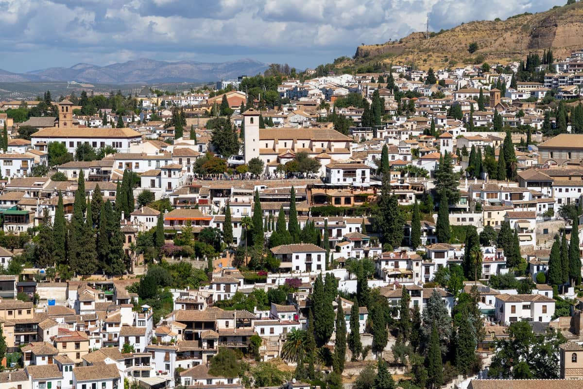วิวทิวทัศน์กว้างของย่าน Sacromonte บนเนินเขาในเมือง Granada พร้อมอาคารสีขาว ต้นไม้สีเขียว และเนินหินในพื้นหลัง