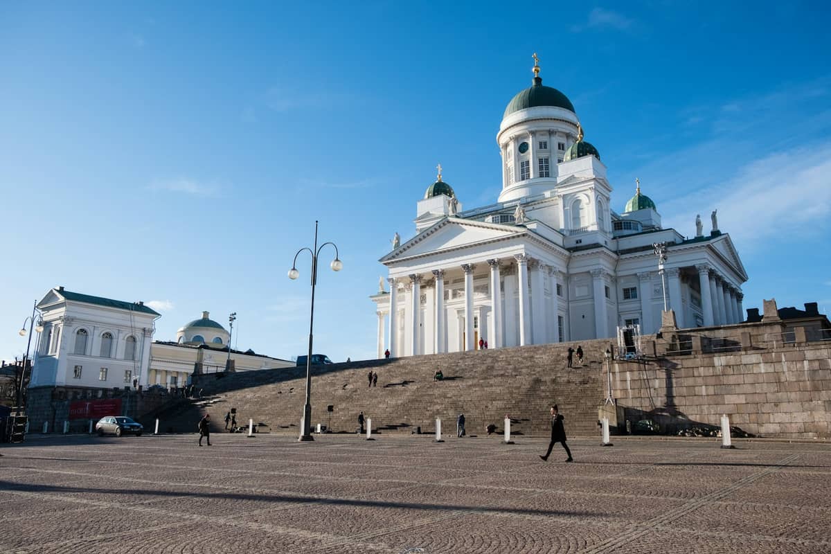 Catedral de Helsinque na Praça do Senado com arquitetura neoclássica, praça de paralelepípedos, amplos degraus e algumas pessoas caminhando sob um céu azul claro.