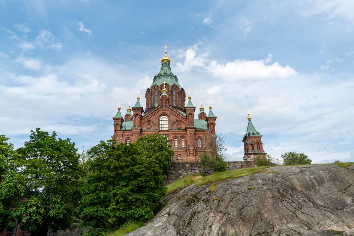 A Catedral Uspenski de tijolo vermelho com cúpulas verdes, vista atrás de rochas e árvores sob um céu parcialmente nublado em Helsinki.