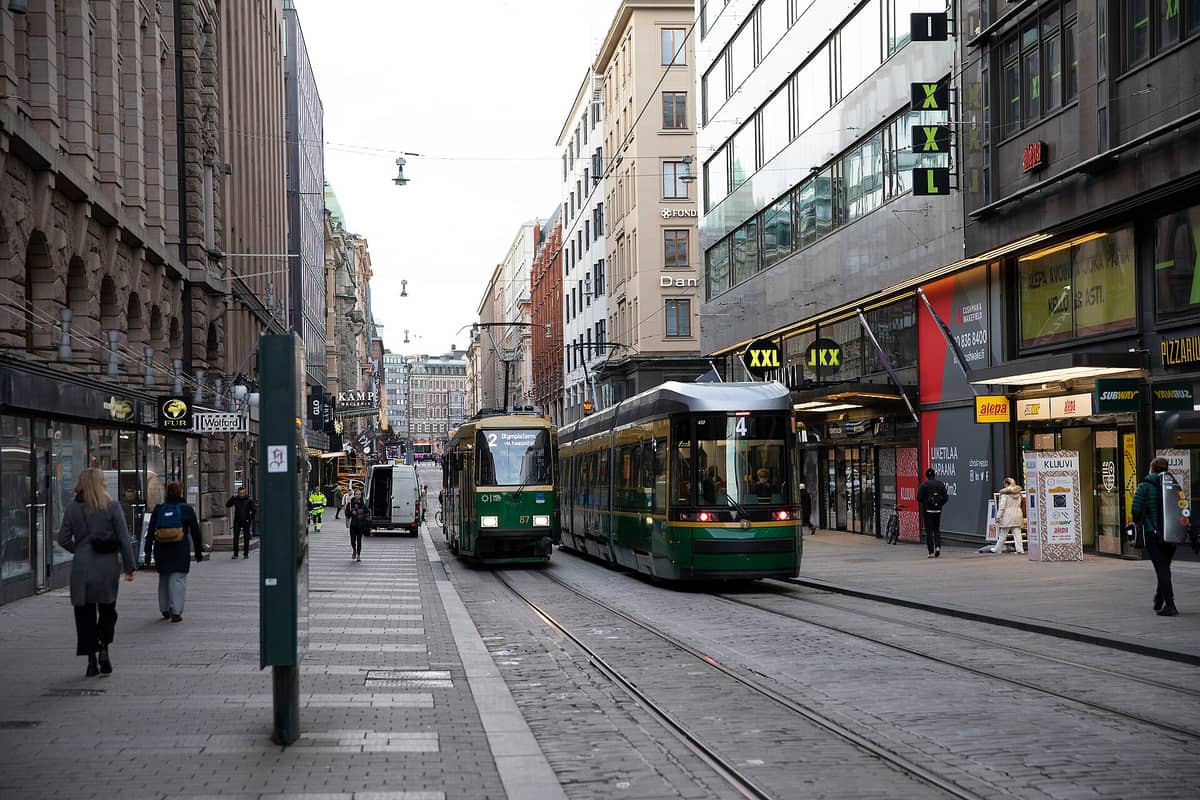 Dois elétricos verdes percorrem uma movimentada rua do centro de Helsínquia, ladeada por lojas e pessoas caminhando nas calçadas.