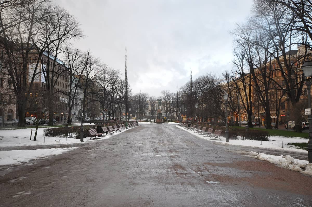 Um amplo bulevar pedestre no centro de Helsinki com bancos, árvores sem folhas e manchas de neve derretendo no inverno. Edifícios da cidade ladeiam ambos os lados.