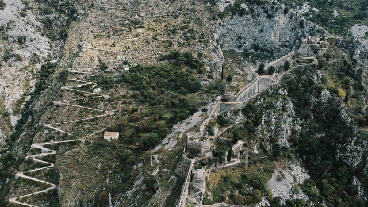 Aerial view of Kotor’s fortress wall zigzagging up the mountain, with ruins and dramatic rocky scenery clearly visible.