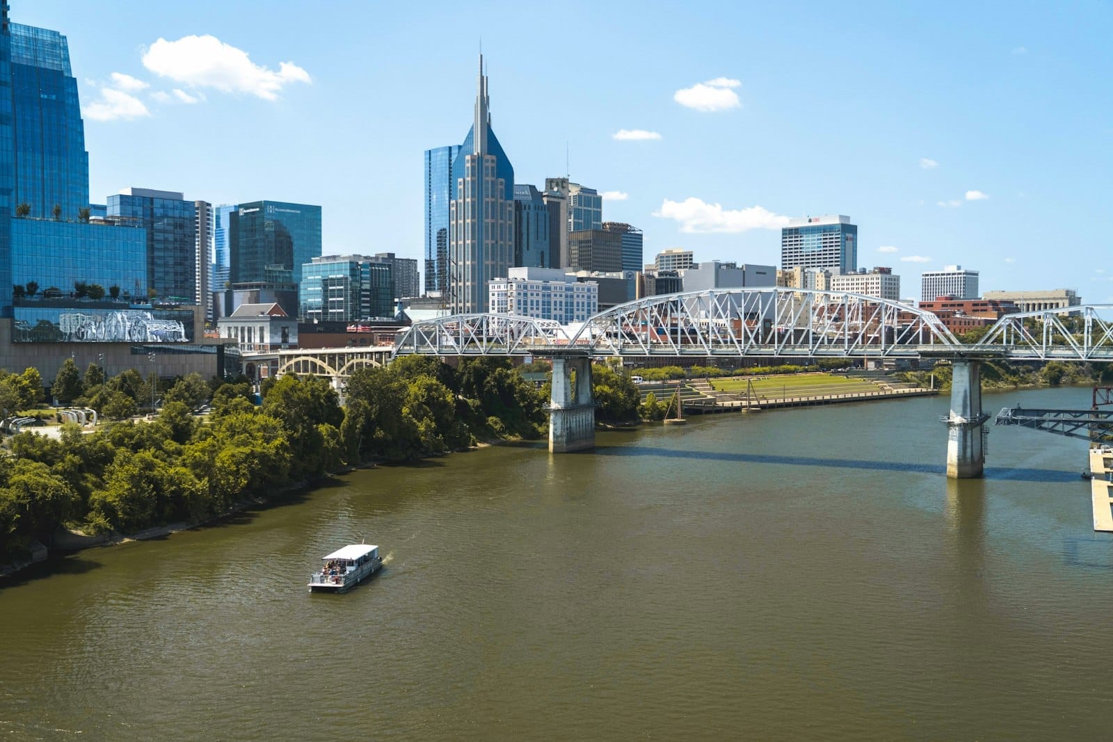 Vista panorámica del horizonte de Nashville con el edificio Batman, rascacielos modernos, un puente peatonal, el río Cumberland y un barco navegando bajo un cielo azul despejado.