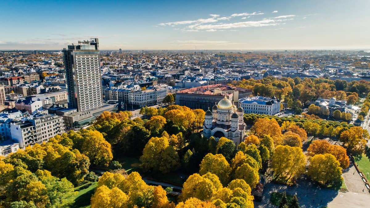 Luftaufnahme von Riga mit bunten Herbstbäumen, Stadtgebäuden und der goldenen Kuppel der Geburtskathedrale unter einem klaren blauen Himmel.