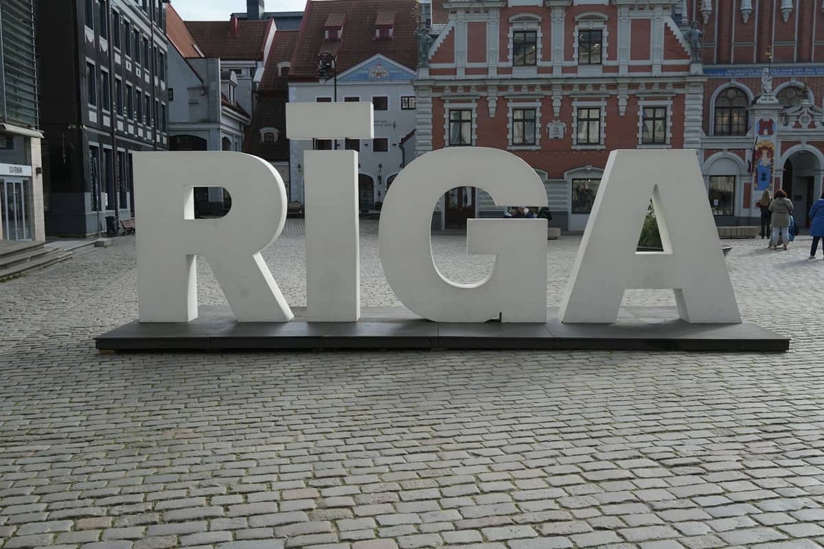 Großes weißes RIGA-Schild auf einem Kopfsteinpflasterplatz mit historischen, bunten Gebäuden der Altstadt von Riga, Lettland, im Hintergrund.
