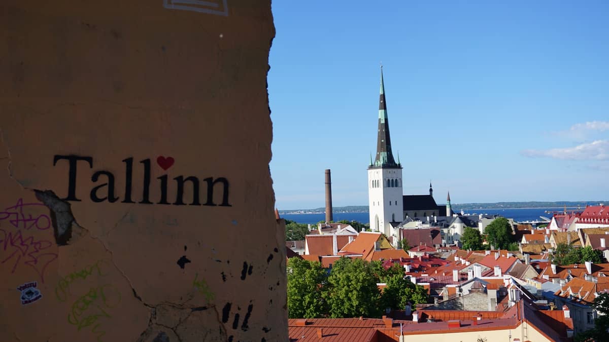 Blick über die Tallinner Altstadt mit dem Schriftzug 'Tallinn' an einer Mauer im Vordergrund und dem Turm der Olaikirche, der hoch über die mittelalterliche Stadtsilhouette ragt.