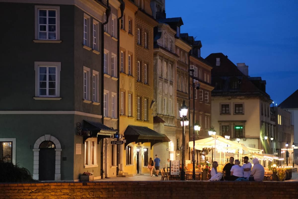 Wieczorna scena uliczna w Warszawie z kolorowymi historycznymi budynkami, restauracjami na świeżym powietrzu, ciepłym oświetleniem i ludźmi jedzącymi kolację pod markizami.