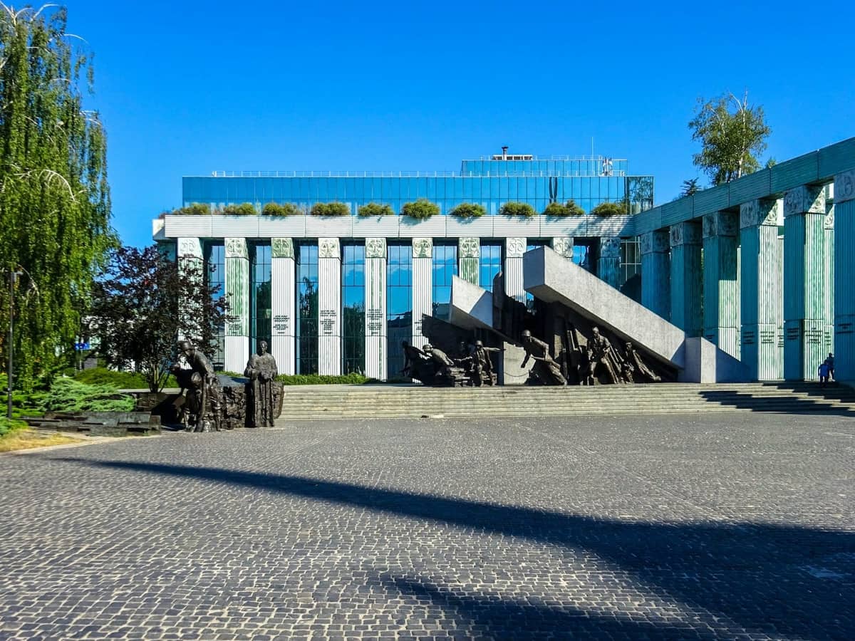 Nowoczesny budynek z dużymi kolumnami i Pomnikiem Powstania Warszawskiego na pierwszym planie, pod bezchmurnym błękitnym niebem.