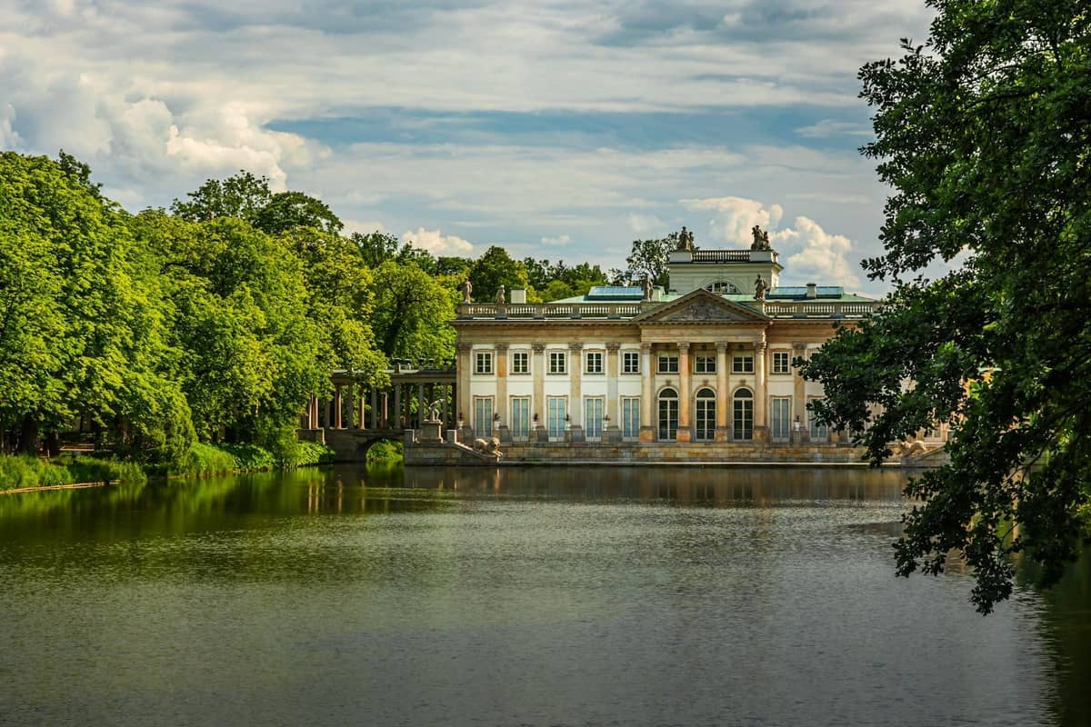 Pałac na Wyspie w warszawskim Parku Łazienki, widoczny ponad spokojnym jeziorem otoczonym bujnymi zielonymi drzewami pod pochmurnym niebem.