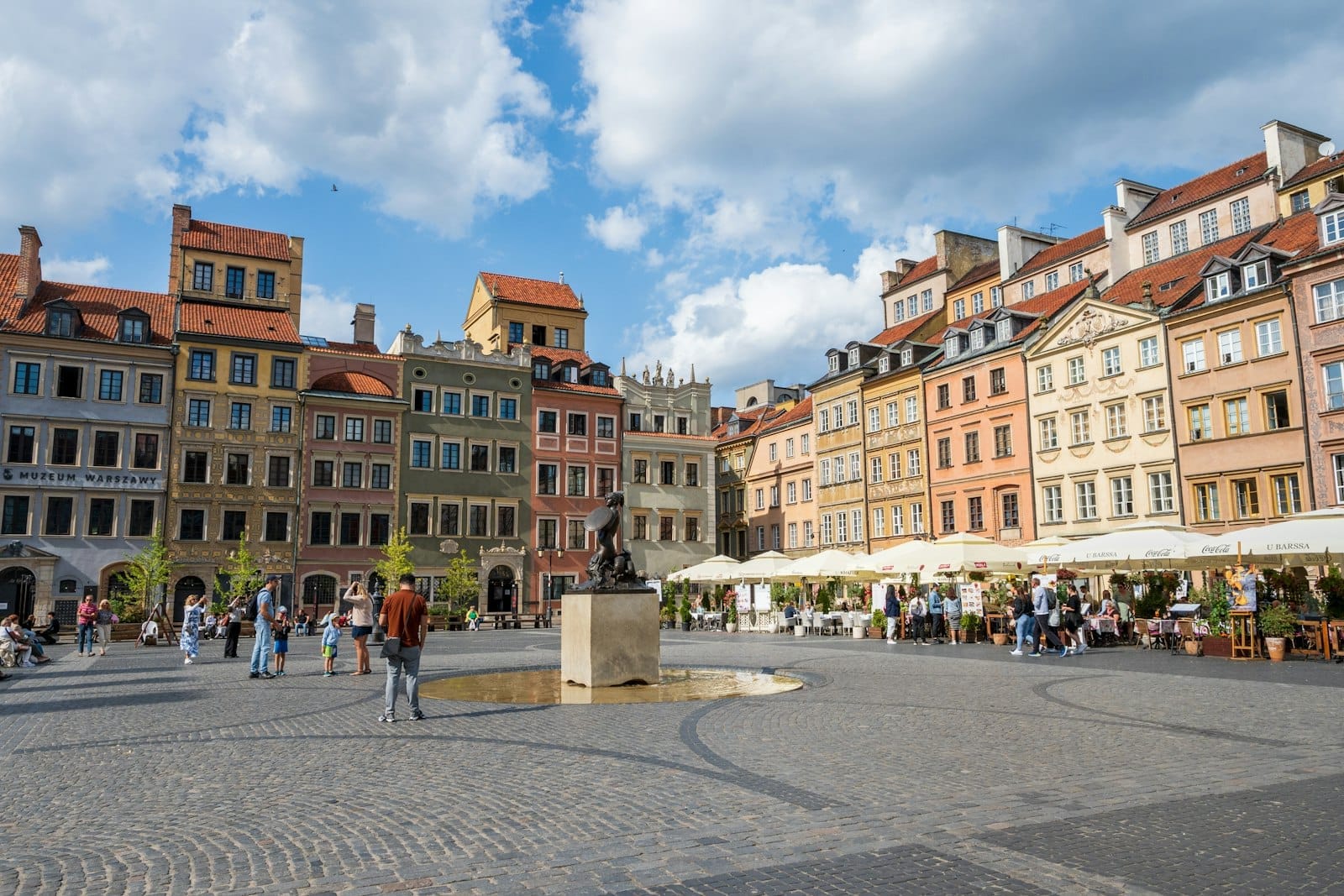 Vista ampla da Praça da Cidade Velha de Varsóvia em um dia ensolarado com edifícios históricos coloridos, a Coluna de Sigismundo, pessoas caminhando e céu azul com nuvens.