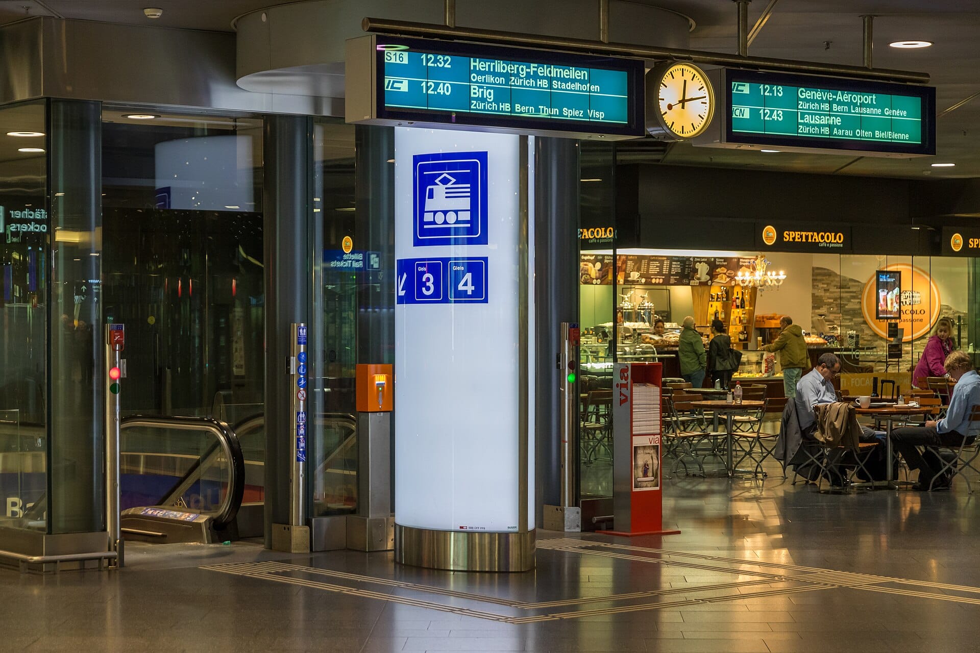 ทางเข้าสถานีรถไฟของสนามบิน Zurich พร้อมบันไดเลื่อน ไอคอนรถไฟ ป้ายแสดงเวลาออกเดินทาง และร้านกาแฟในพื้นหลัง