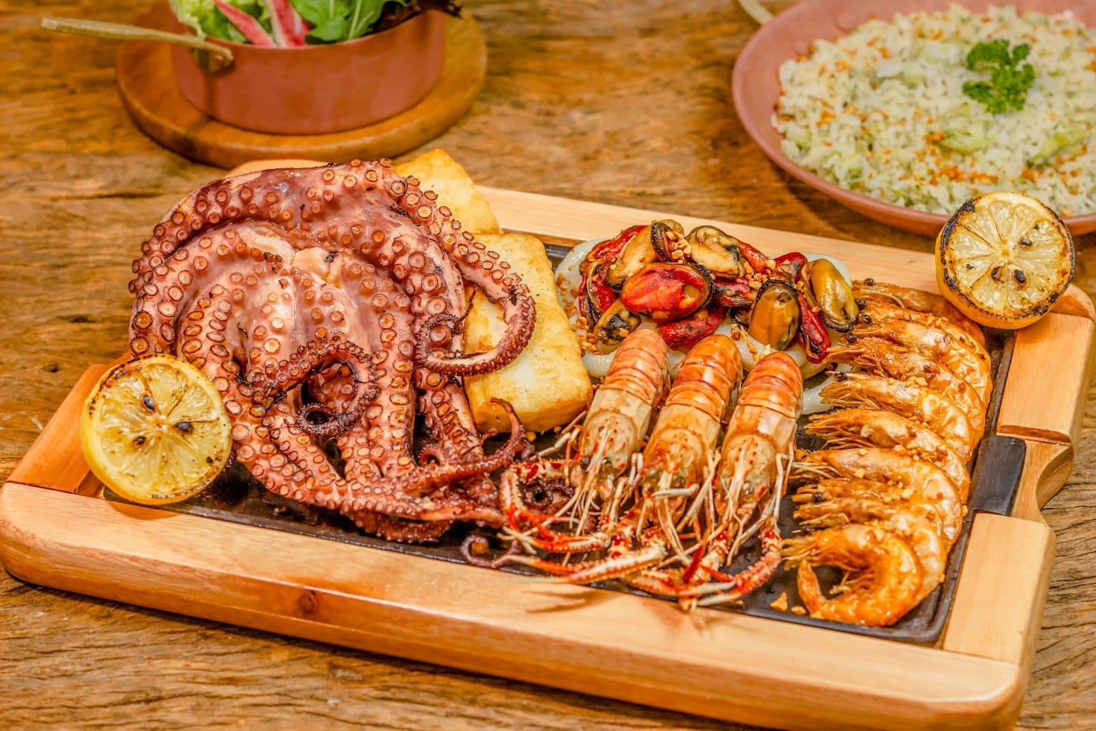 Eine Platte mit frisch gegrillten Meeresfrüchten, darunter Tintenfisch, Garnelen, Muscheln und Zitrone, serviert auf einem Holzbrett in einem rustikalen Restaurant.