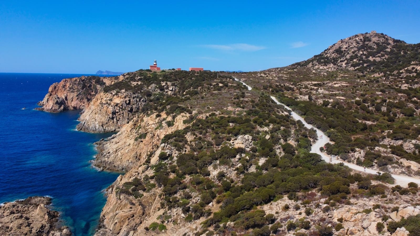 Felsige Küstenlandschaft mit einer kurvenreichen Straße und einem Leuchtturm in der Ferne an einem sonnigen Tag auf Sardinien