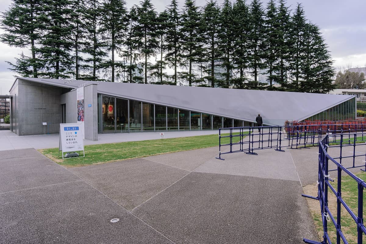 Exterior view of 21_21 Design Sight museum in Tokyo with its distinctive modern sloped roof and glass facade, nestled next to a row of tall trees.