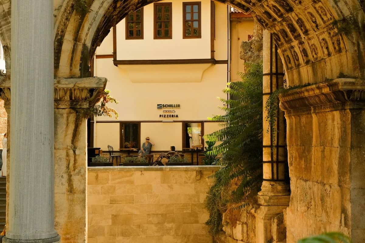 View through the stone arches of Hadrian’s Gate toward an Ottoman-era house in Antalya’s Kaleiçi old town on a sunny day.