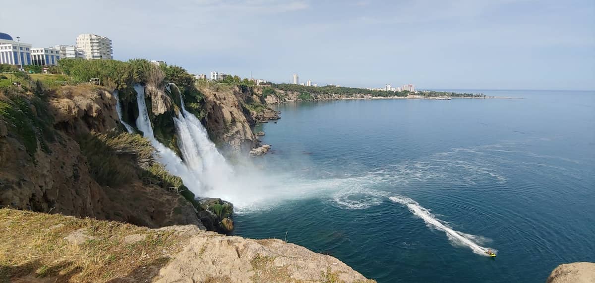 Wide view of Düden Waterfalls cascading from cliffs into the sea with Antalya city buildings and a boat on the water.