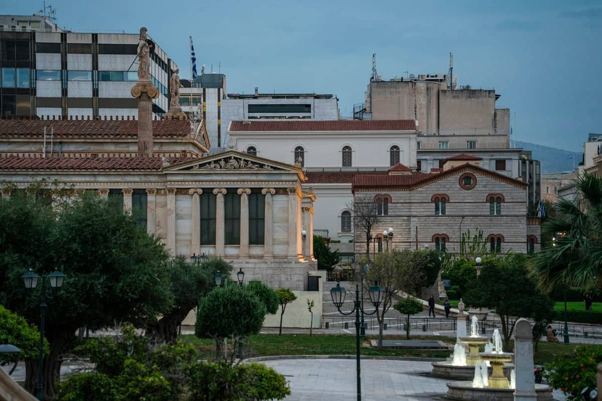 Neoklassizistische Gebäude und Statue auf einem Platz in einem historischen Viertel in Athen, umgeben von Stadtarchitektur und Grün in der Abenddämmerung.