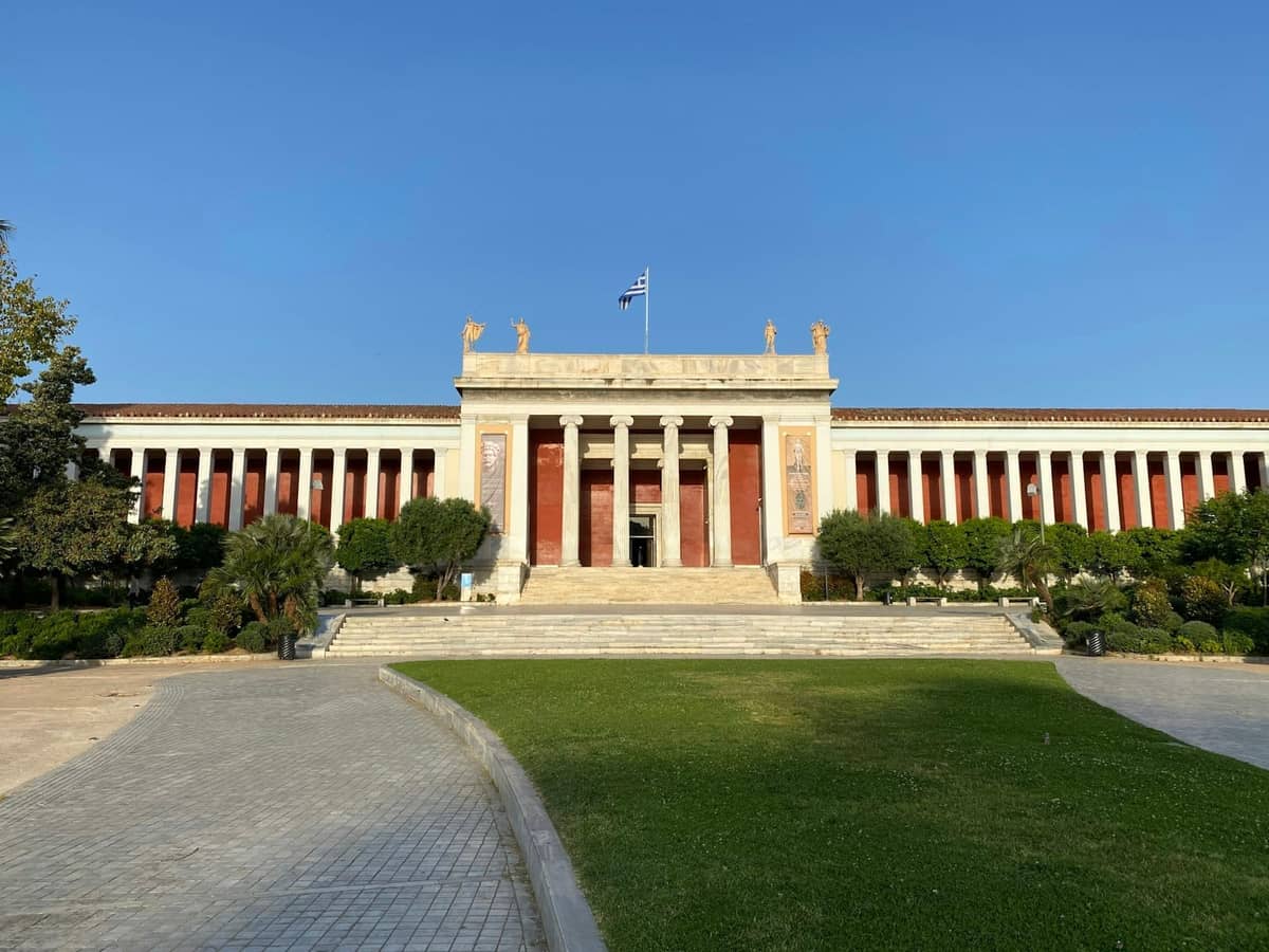 Weitwinkelansicht des Nationalen Archäologischen Museums in Athen, mit neoklassischen Säulen, Statuen, griechischer Flagge und gepflegtem Garten davor.