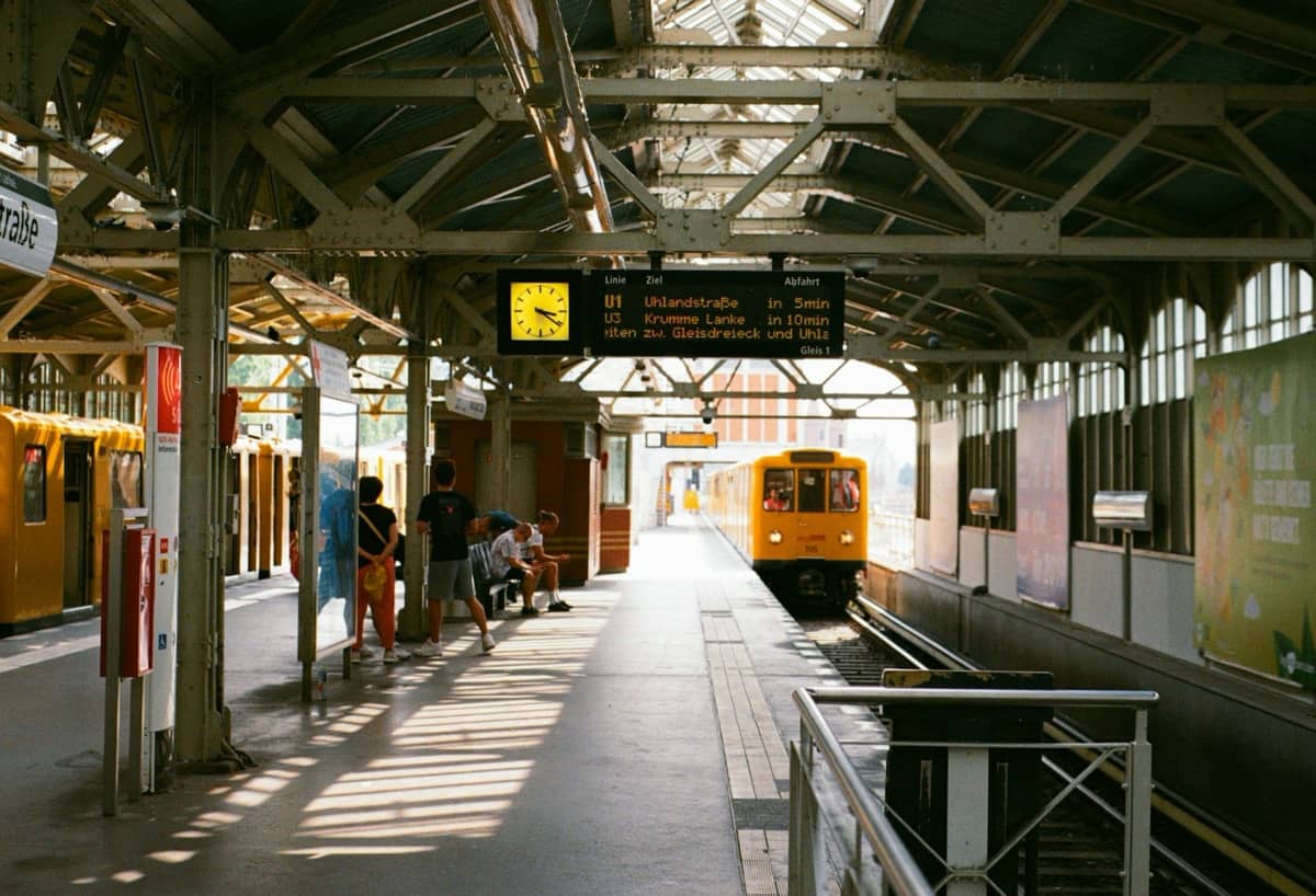 Estación de U-Bahn de Berlín con tren amarillo llegando, personas esperando en el andén cubierto y panel de horarios en lo alto.