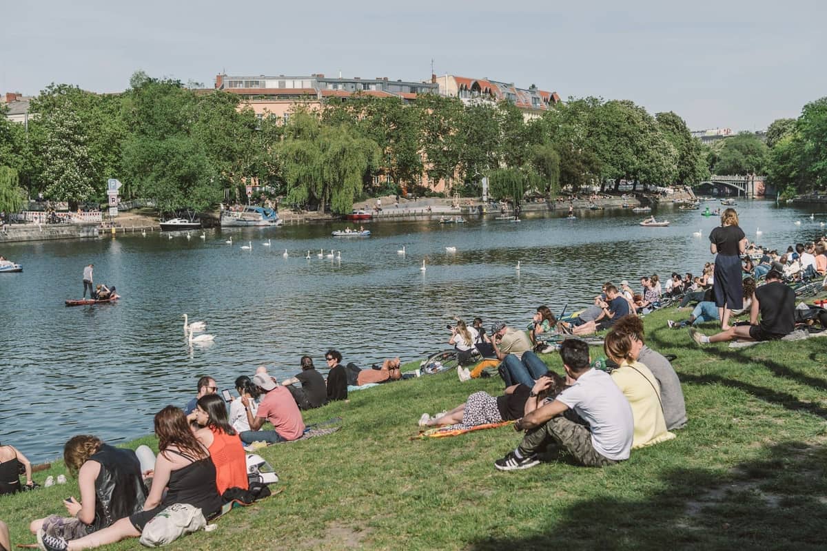 Personas descansando en el césped junto a un río en Berlín en un día soleado, con cisnes en el agua y árboles al fondo.