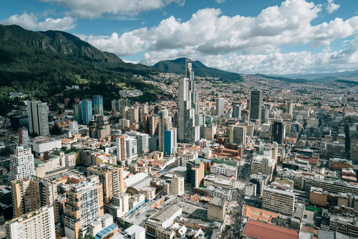 A wide aerial view of Bogotá showing the downtown cityscape, tall modern buildings, mountainous background, and dense urban sprawl under a partly cloudy sky.