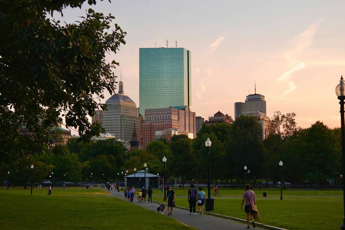 Boston Common o zachodzie słońca z panoramą miasta w tle, ludzie spacerujący po ścieżkach, zielone drzewa i otwarta przestrzeń parkowa.