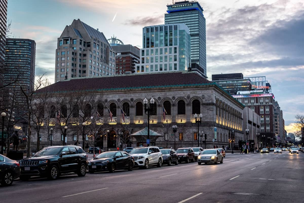 Boston Public Library na Copley Square, z nowoczesnymi wieżowcami Back Bay widocznymi w tle i samochodami ustawionymi wzdłuż ulicy na pierwszym planie.