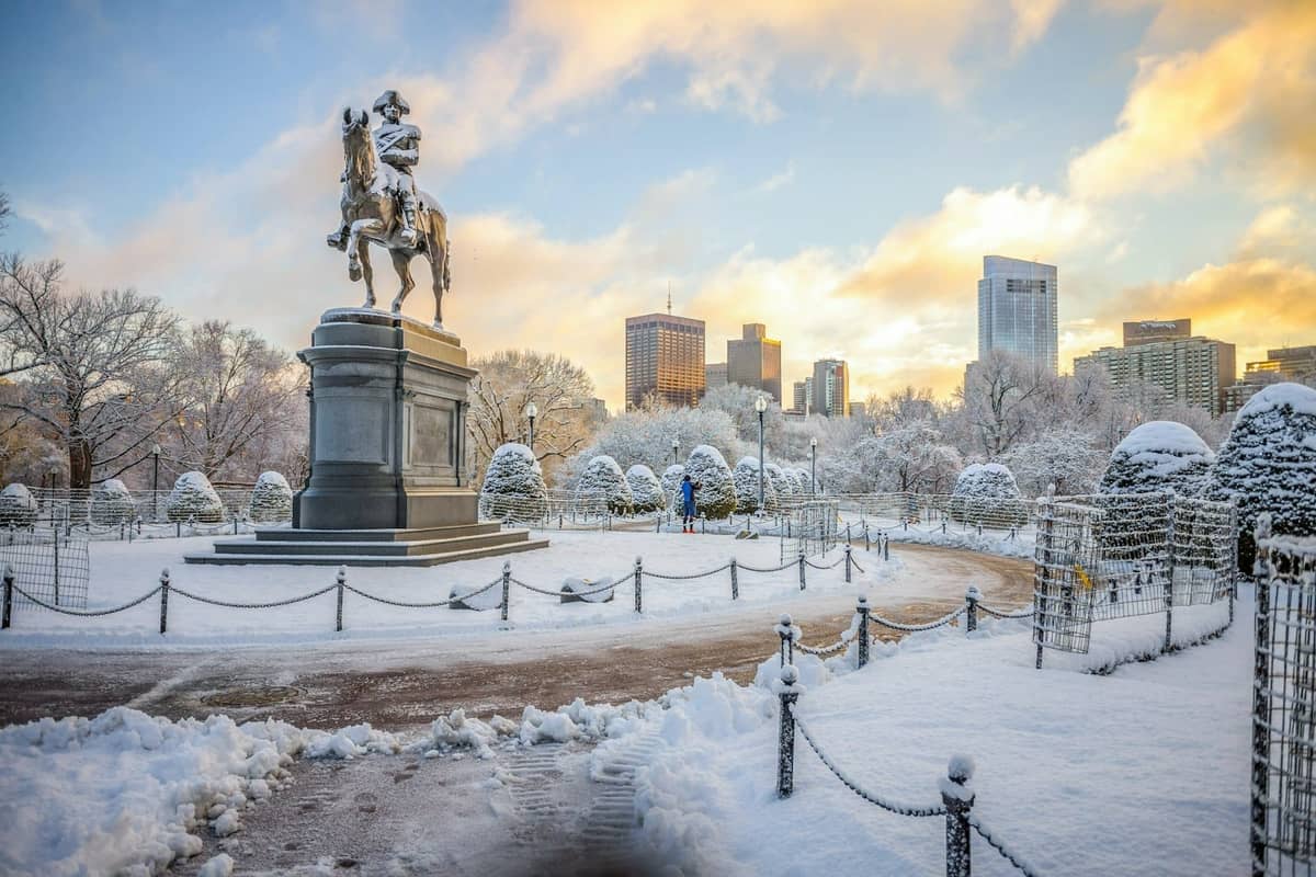 Ośnieżony Boston Public Garden z konną statuą George'a Washingtona, panoramą miasta i zimowym niebem o zachodzie słońca.