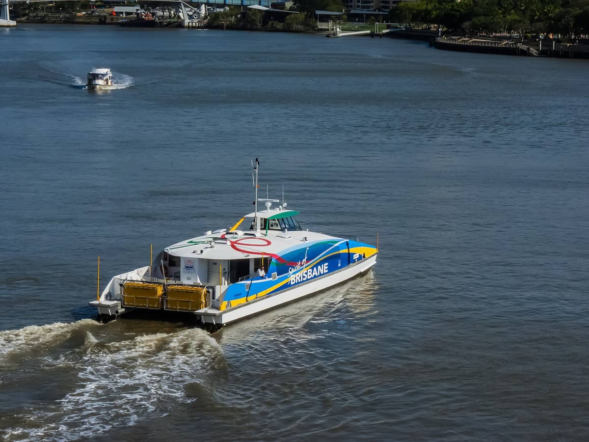 A modern CityCat ferry labeled 'Brisbane' traveling along the Brisbane River, with other ferries and riverside in the background.