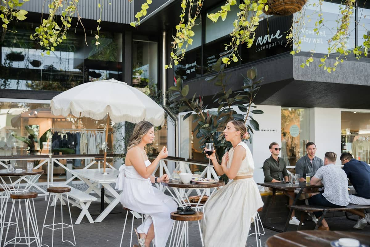 People dining and enjoying drinks at an outdoor café with modern Brisbane architecture and greenery in the background.