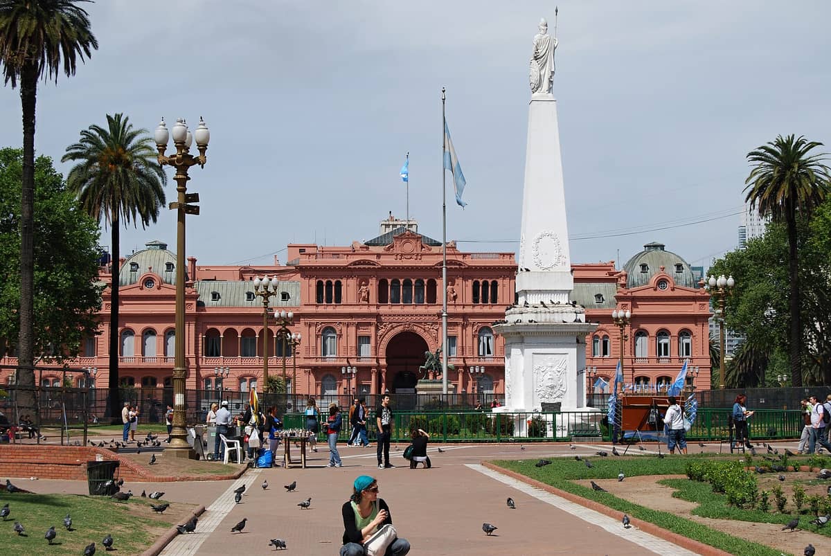 มุมมองกว้างของ Plaza de Mayo ในบัวโนสไอเรส พร้อม Casa Rosada และ Pirámide de Mayo สีขาวที่ล้อมรอบด้วยผู้คนและต้นปาล์ม