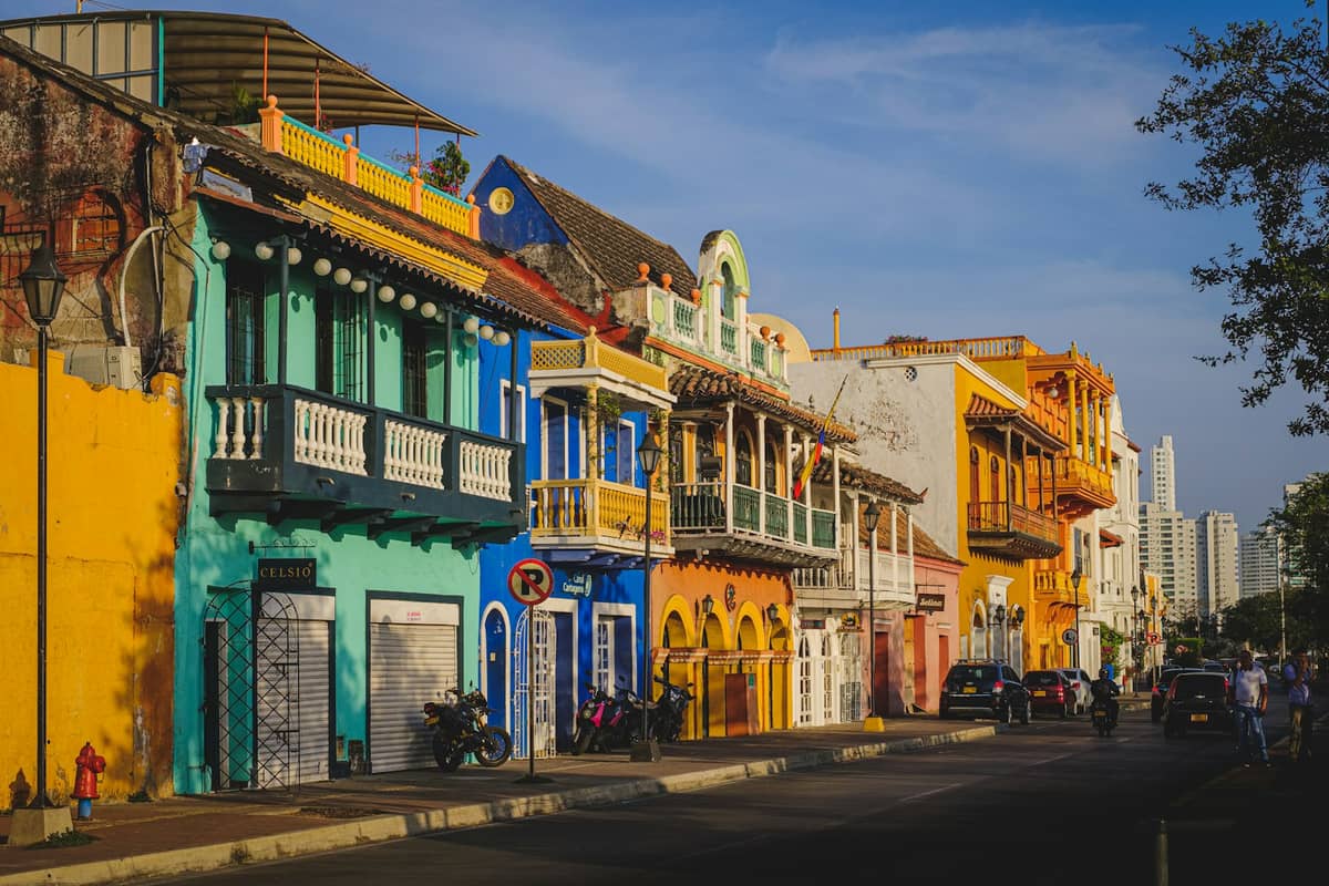 Colorful colonial-style buildings with balconies line a street in Cartagena, Colombia, with people and cars in the afternoon light.