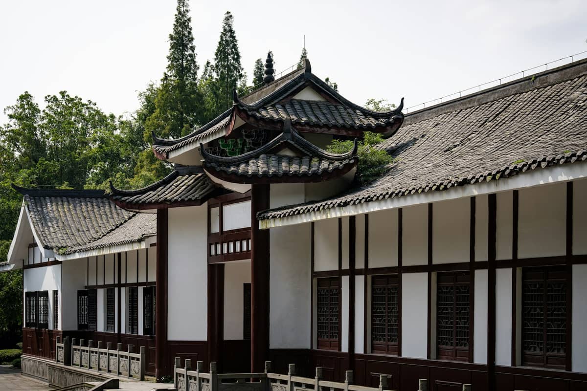 Traditionelles chinesisches Klostergebäude mit weißen Wänden, dunklen Holzdetails, geschwungenen Dachtraufen und Bäumen im Hintergrund an einem sonnigen Tag.