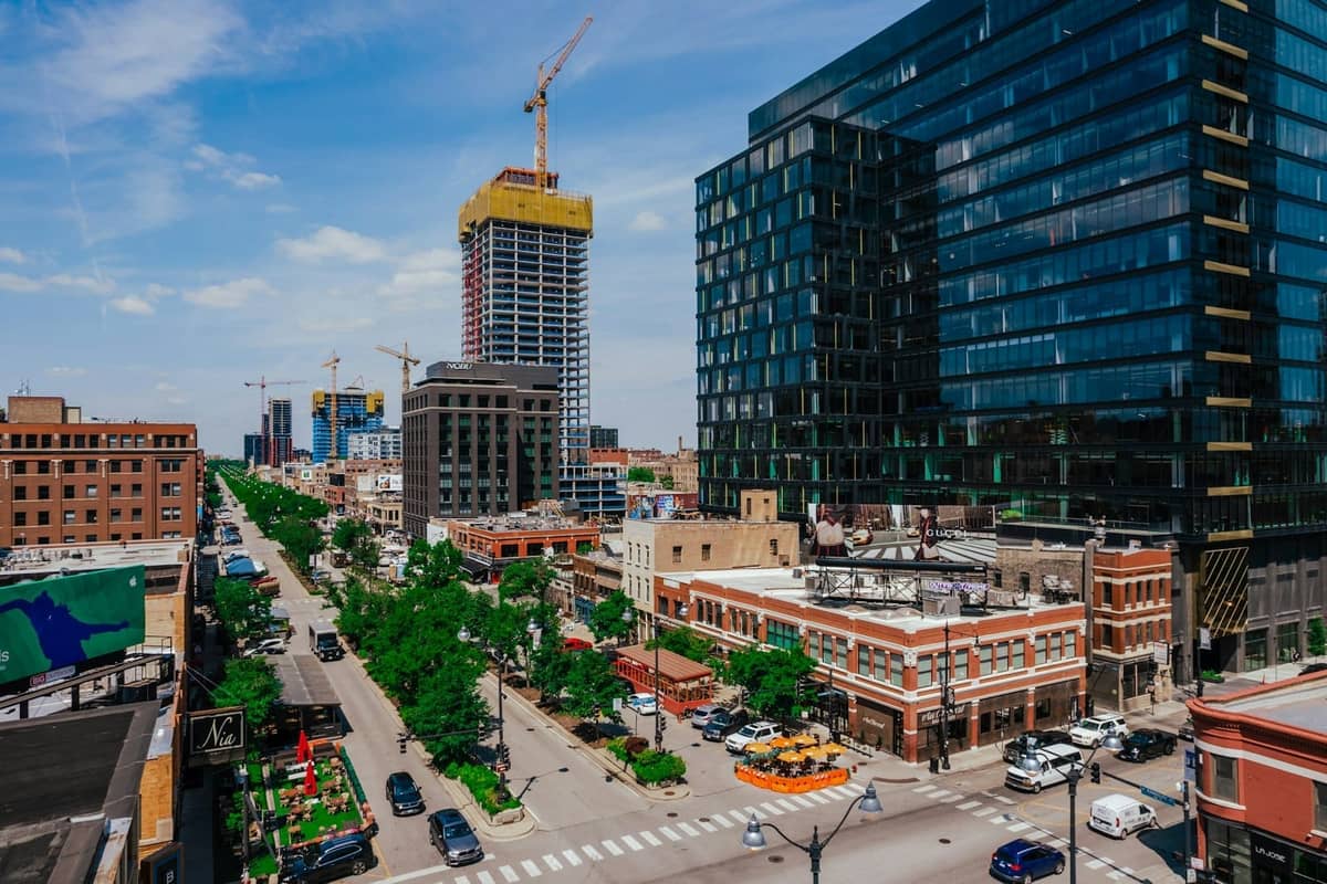 Lebhafte Stadtstraße in einem Chicago-Viertel mit neuen Hochhäusern, baumgesäumten Gehwegen, Restaurants und Kränen im Hintergrund.