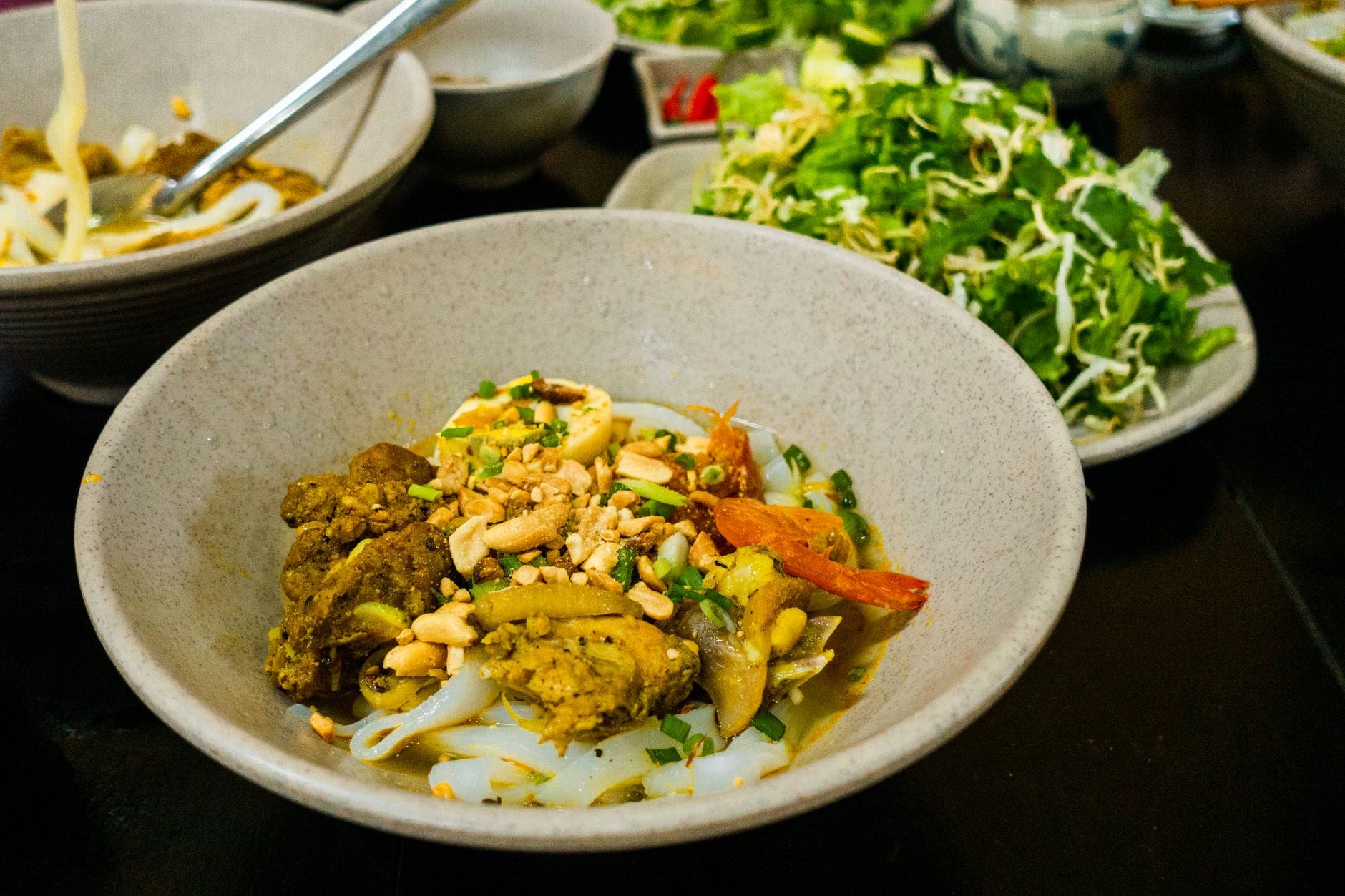ชามมิ๋กว๋าง อาหารเส้นสีเหลืองขมิ้นที่มีหมู กุ้ง ถั่วลิสง และสมุนไพร ล้อมรอบด้วยผักสดบนโต๊ะสีเข้ม