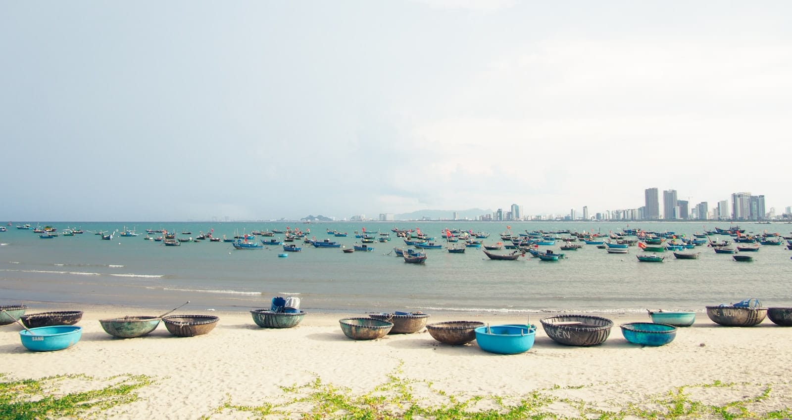 วิวพาโนรามาของชายหาดดานังที่มีเรือกลมบนผืนทราย เรือประมงจำนวนมากในน้ำ และเส้นขอบฟ้าของเมืองในระยะไกล