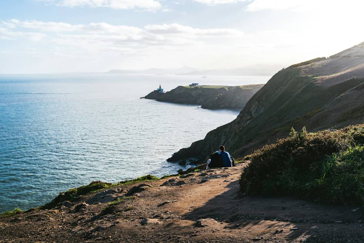 Widok na nadmorskie klify w Howth niedaleko Dublina ze ścieżką spacerową, morzem i latarnią morską w oddali w słoneczny dzień.